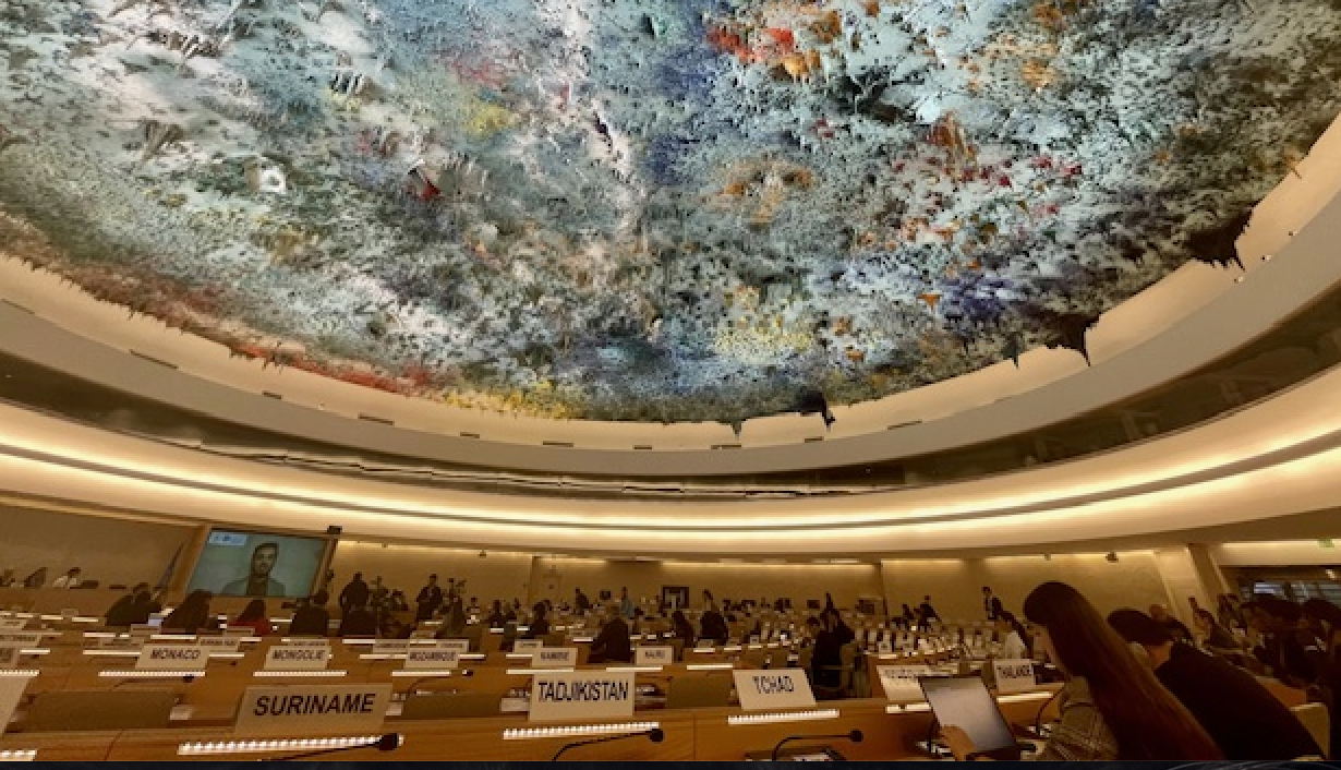 An ornate, colorful ceiling looms over a United Nations meeting room with country nameplates and delegates seated at rows of desks, as others gather and converse throughout the chamber.